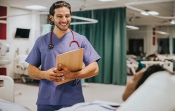 Male nurse holding a clipboard, smiling at a patient in bed. 